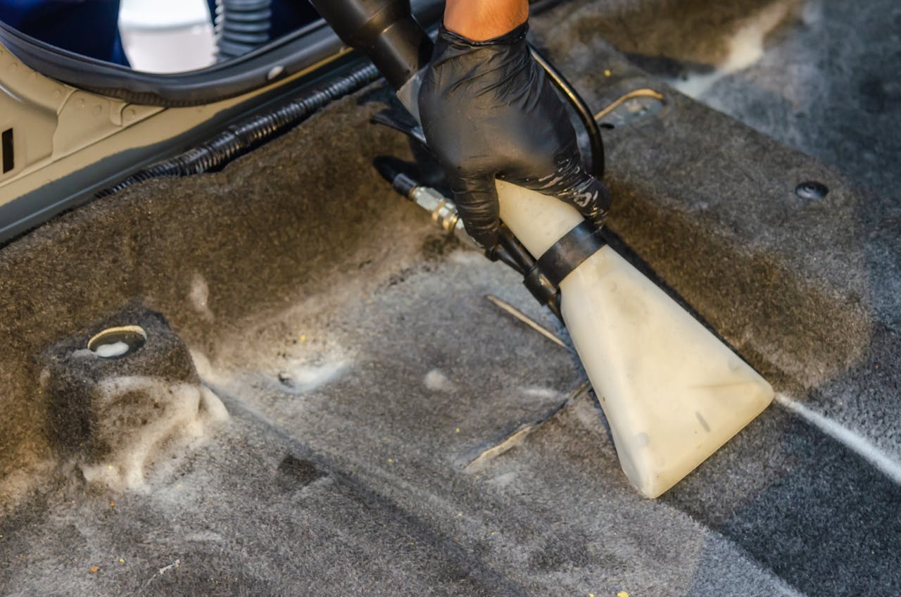 gallery-03 A hand in a glove using a vacuum to clean a car interior carpet, removing dirt effectively.